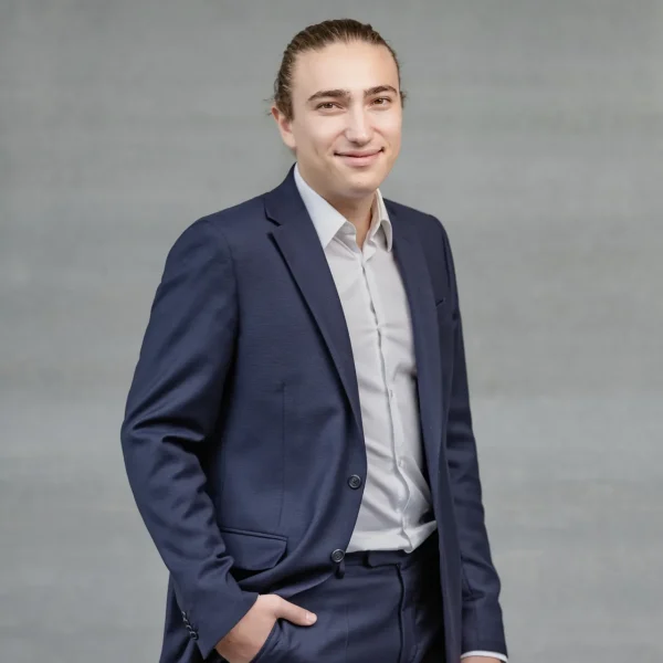 Young man in a navy suit posing with hands in pockets against a gray backdrop, smiling slightly.