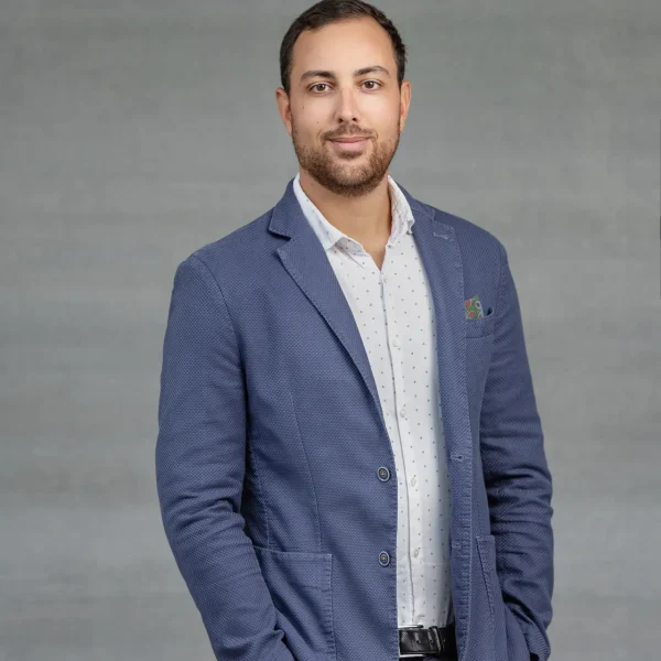 Portrait of a man in a blue blazer and white patterned shirt, hands in pockets, standing against a gray backdrop.