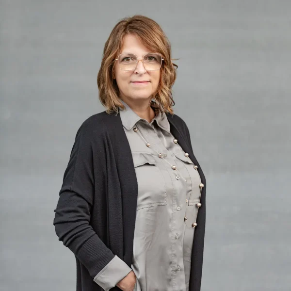 Portrait of a middle-aged woman with glasses, wearing a gray blouse and black cardigan, standing against a gray backdrop.