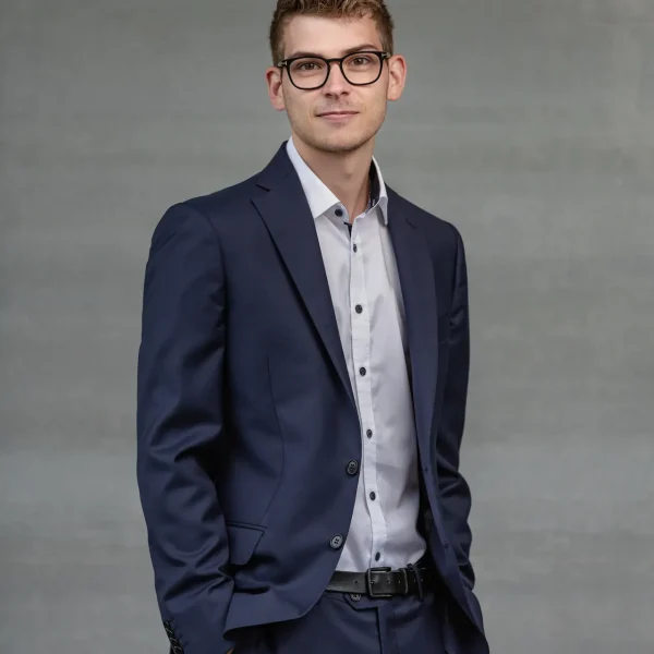 Professional head-and-shoulders portrait of a young man in a navy suit, white shirt, and glasses, posing against a gray backdrop.