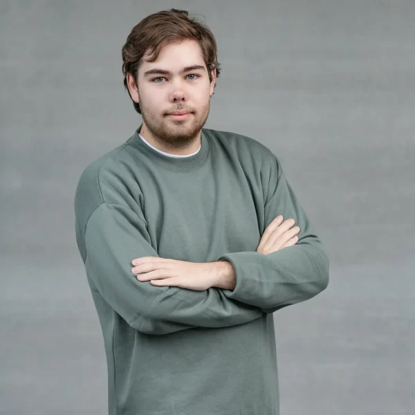 Young man with light facial hair wearing a green sweatshirt, arms crossed, looking at the camera against a gray backdrop.