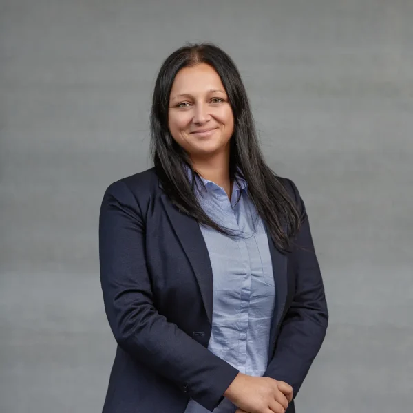 Portrait of a woman in a navy blazer and light blue shirt smiling against a gray studio backdrop.