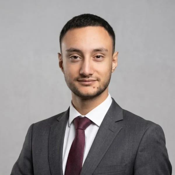 Professional headshot: man in a gray suit, white shirt, burgundy tie, smiling slightly against a neutral gray background.