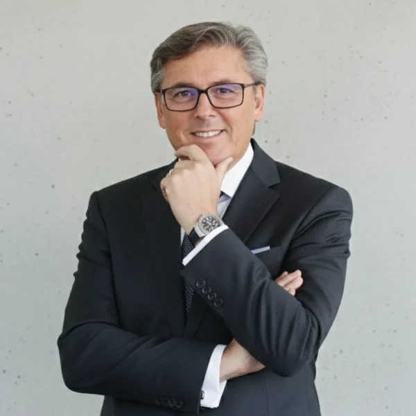 Professional man in a dark suit and glasses posing with his hand on his chin against a light gray wall backdrop.