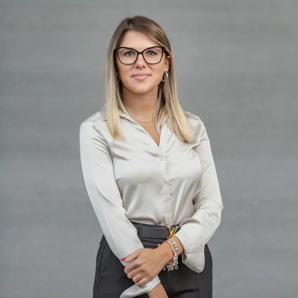 Professional woman with glasses posing against a gray background, wearing a silver blouse and black pants, arms loosely crossed.