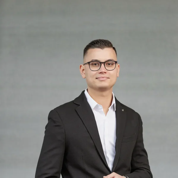 Young man in a black blazer and white shirt, posing with one hand in his pocket against a gray backdrop, wearing glasses and a wristwatch.
