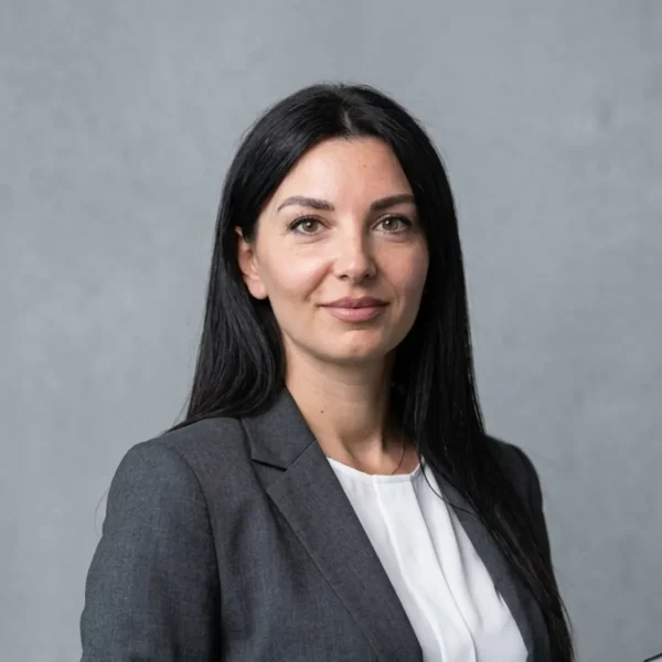 Professional headshot of a woman with long dark hair, wearing a gray blazer and white blouse, against a neutral gray backdrop.