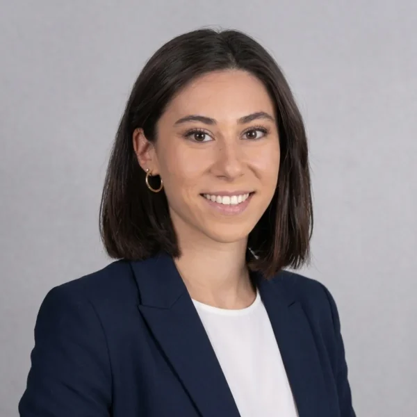 Professional headshot of a woman with shoulder-length dark hair, wearing a navy blazer and white top, smiling against a gray backdrop 2C/ neutral background.
