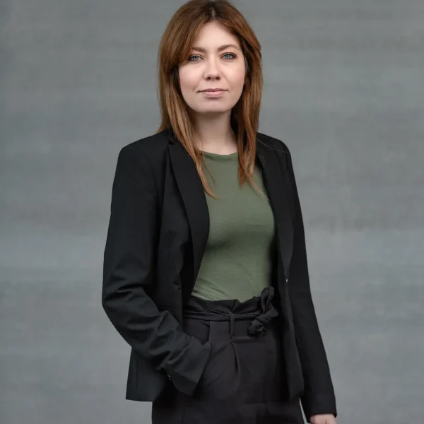 Portrait of a woman with shoulder-length brown hair in a black blazer and green top, standing against a gray wall.
