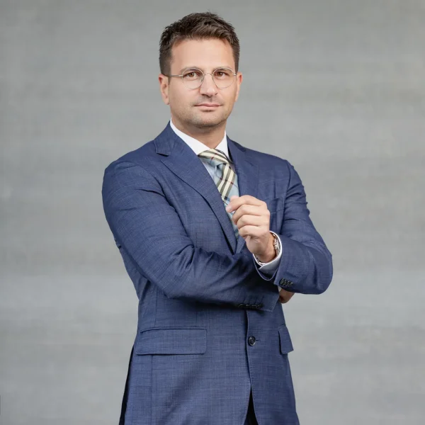 Man in a blue suit with arms crossed, facing the camera against a gray studio backdrop.