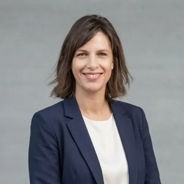 Professional headshot: smiling woman with shoulder-length brown hair in a navy blazer against a gray background.