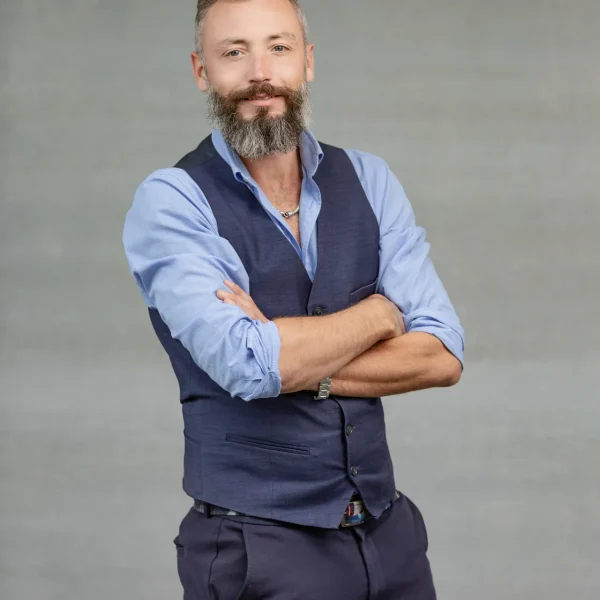 Professional man with a gray beard in a blue dress shirt and navy vest, arms crossed against a neutral gray backdrop.