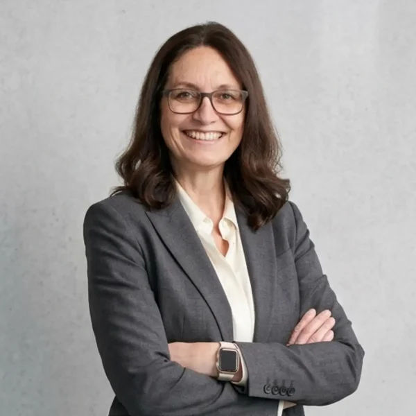 Professional woman with shoulder-length brown hair and glasses, wearing a gray blazer and white blouse, smiling with arms crossed against a light gray background.