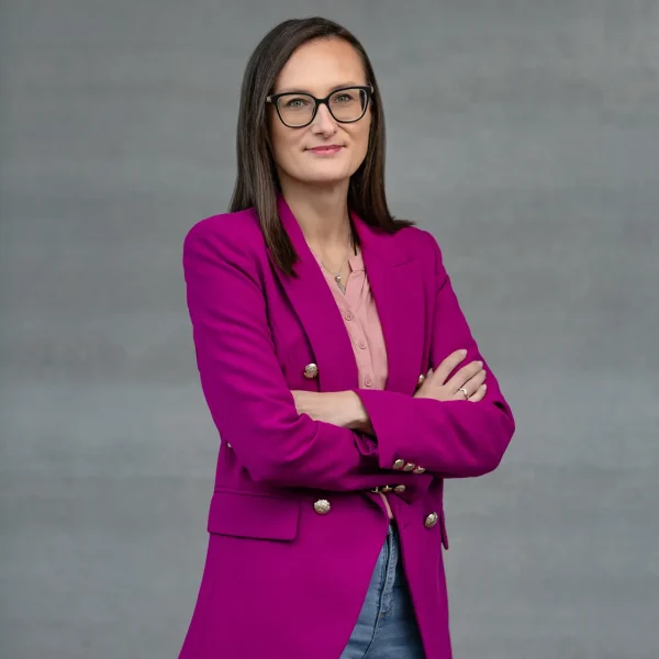 Woman in a magenta blazer and pink blouse stands with arms crossed, looking at the camera against a gray backdrop.