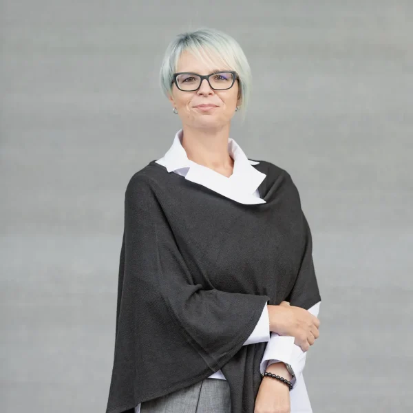 Smiling woman with short light-gray hair and glasses, wearing a black poncho over a white-collared shirt against a gray backdrop.